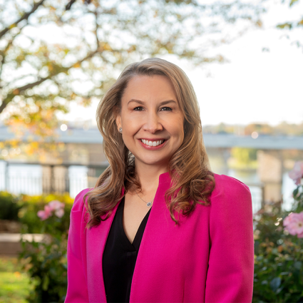 A smiling Caucasian female-presenting individual with blonde hair wearing a bright pink suit jacket against a nature-like background