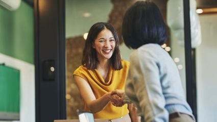 Two people in a professional setting smiling and shaking hands, symbolizing agreement or partnership.