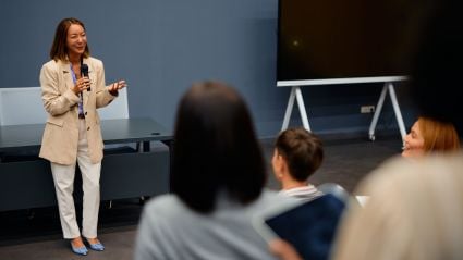 Professional woman holding a microphone while speaking to an audience in a business setting.