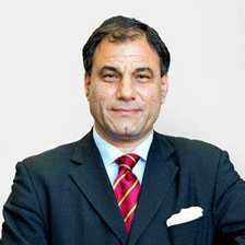 Lord Karan Bilimoria, dressed in a dark suit with a red and gold striped tie and white pocket square, poses confidently against a light background. He is the founder of Cobra Beer, a CBE and Deputy Lieutenant of Greater London, and a prominent figure in UK-India business relations.