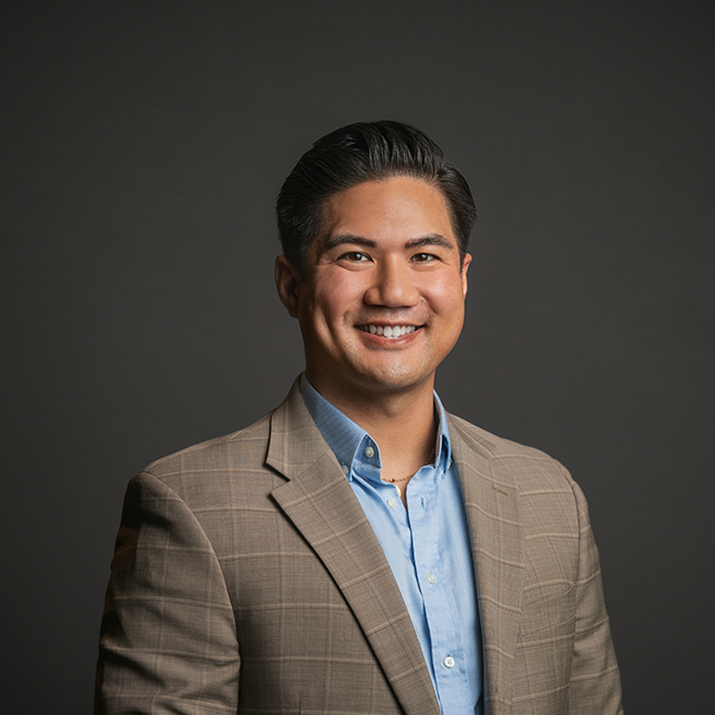 Professional headshot of Brian Carandang, a man with neatly styled black hair, wearing a beige plaid blazer over a light blue button-down shirt. He is smiling and posed against a dark gray background.