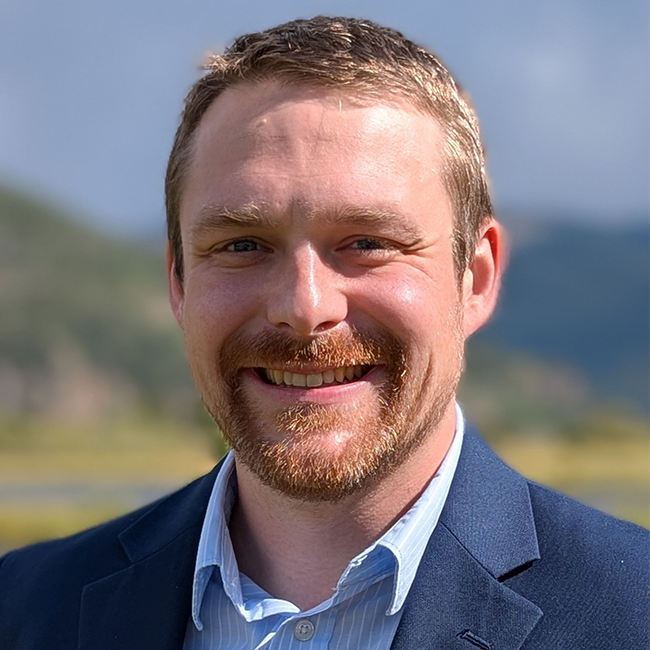 Headshot of a smiling man with short light brown hair and a trimmed beard, wearing a blue blazer and light blue collared shirt, outdoors with blurred greenery and mountains in the background.