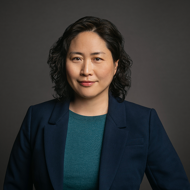Professional headshot of June Cha, a woman with short curly black hair, wearing a teal top and navy blazer. She is posed against a dark gray background with a confident expression.