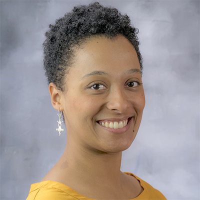 Headshot of a smiling woman with short curly hair, wearing a mustard yellow top and dangling earrings, set against a soft gray studio backdrop.
