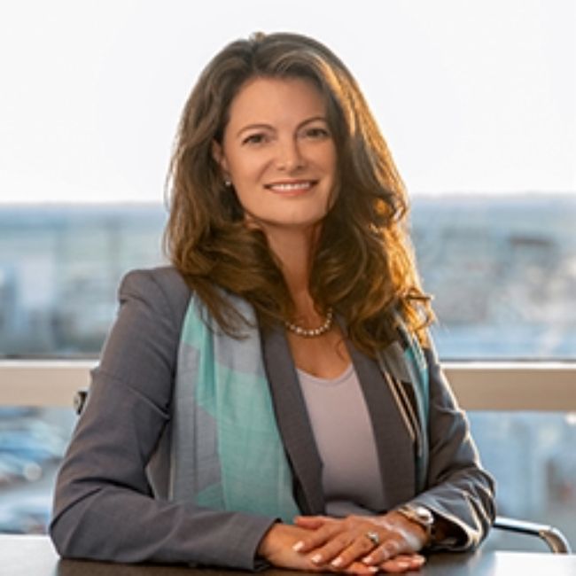 Kristin Barens, a woman with long brown hair, wearing a gray blazer over a light top with a patterned scarf and a pearl necklace, seated at a desk and smiling, with a blurred cityscape in the background.