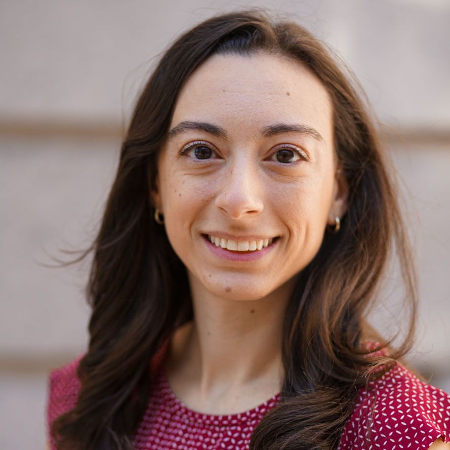 A woman with long dark hair smiles at the camera, wearing a red patterned blouse, standing outdoors in natural light.