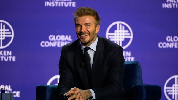 An image of a man named David Beckham, smiling and sitting onstage at the Milken Institute's Global Conference 2024 against a Milken Institute Global Conference blue backdrop. He is wearing a black suit and black tie, and has white skin, a short brunette beard, and short brunette hair.