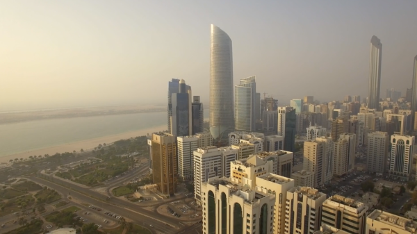 A hazy aerial view of Abu Dhabi’s skyline with modern high-rise towers, including a tall curved glass skyscraper at the center. The city’s grid of roads and beige residential buildings stretch outward, while the coastline with sandy beaches and calm waters appears to the left.