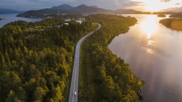 A winding road runs through a dense green forest beside a calm body of water, with cars traveling along it at sunset. The golden light reflects off the water, and distant mountains rise under a partly cloudy sky.