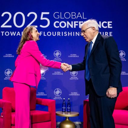 A woman in a bright pink suit and a man in a dark suit shake hands on stage at the 2025 Global Conference. They stand in front of a blue backdrop with Milken Institute and Global Conference logos, with two red chairs and water bottles visible between them.