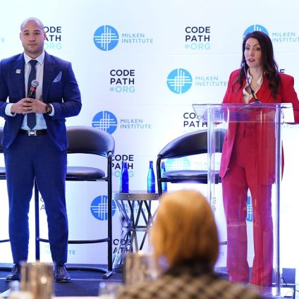 Two speakers stand on stage at a Milken Institute and CodePath-branded event. A woman at a clear podium speaks while holding notes, and a man beside her holds a microphone. Audience members are visible in the foreground.