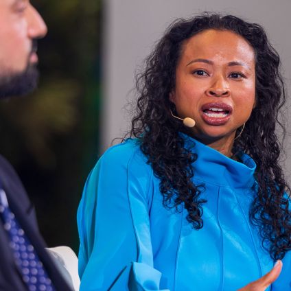 A panelist wearing a bright blue top speaks into a headset microphone during a session. Another panelist is partially visible to the left, slightly out of focus.