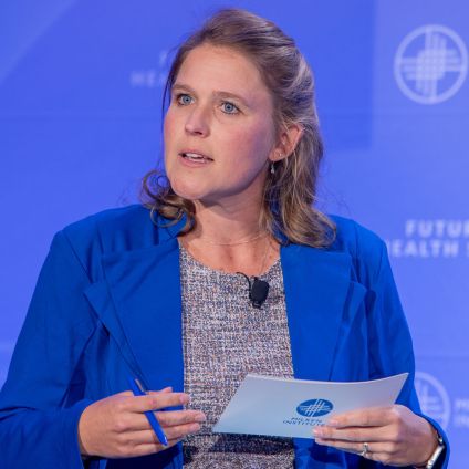 A woman in a royal-blue blazer speaks during a session at the Future of Health Summit. She holds Milken Institute note cards and a pen, with a blue event backdrop behind her.