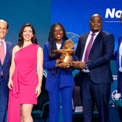 A group of five people stand on stage at the Global Conference during an award presentation. The person in the center holds a large gold-toned award sculpture, with Milken Institute branding visible on the backdrop.