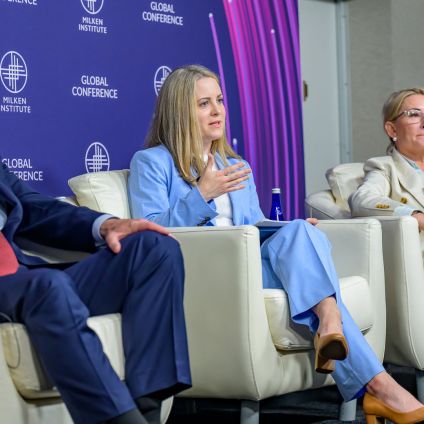 Three panelists sit in white armchairs onstage at a conference, speaking in front of a blue Milken Institute Global Conference backdrop. A man in a dark suit and red tie sits on the left, a woman in a light blue suit speaks while gesturing with one hand in the center, and another woman in a beige blazer and tan pants sits on the right holding a notepad and pen.