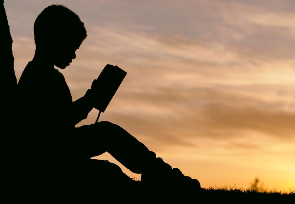 A child's silhouette sitting down reading a book against a sunset