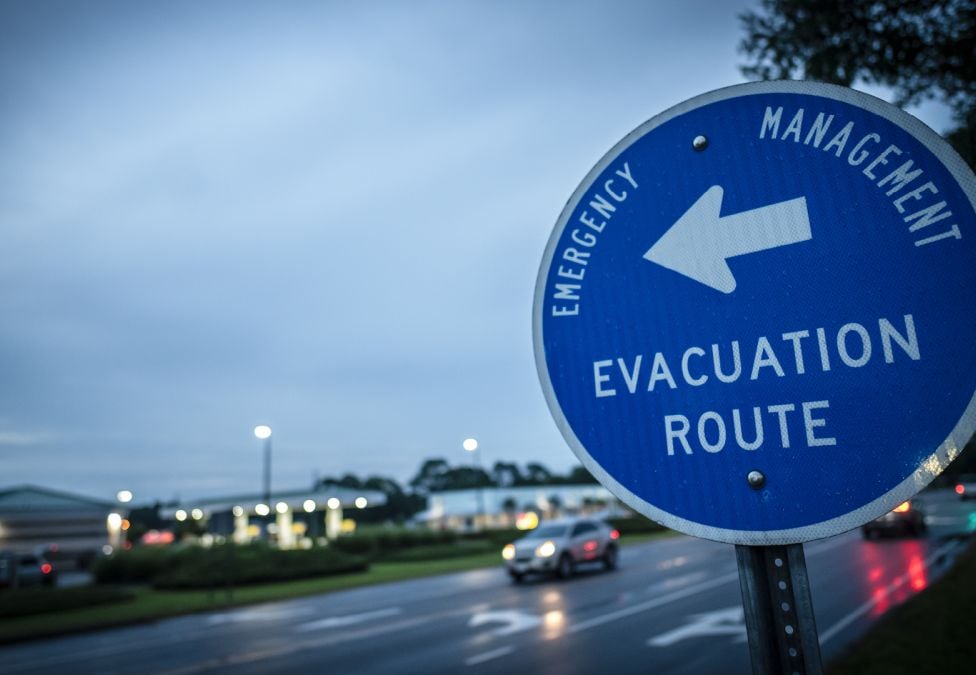 Blue emergency management sign showing an evacuation route with a left-pointing arrow, positioned along a wet roadway at dusk with cars and buildings in the background.