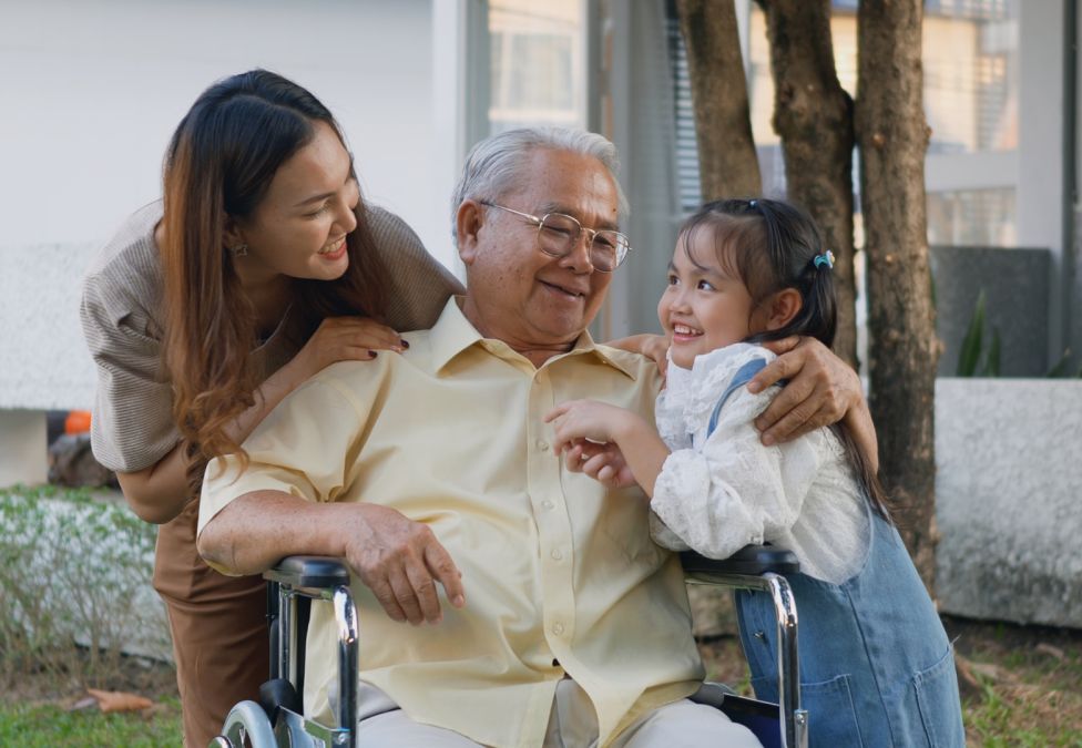 An elderly man in a wheelchair smiles warmly as he is embraced by a young woman and a little girl outdoors, sharing a joyful family moment together.