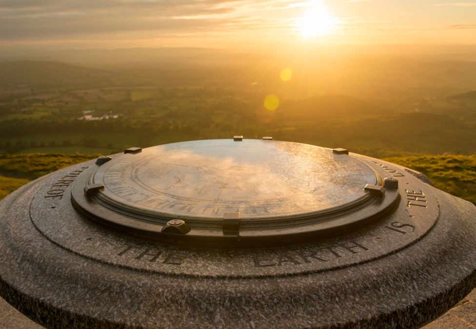 Stone compass viewpoint on a hilltop at sunset, with golden sunlight casting a warm glow over the engraved circular map and the rolling countryside landscape in the distance.