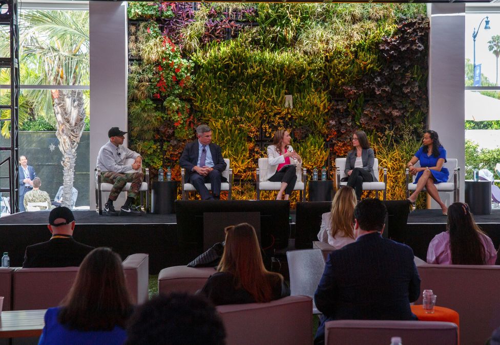 A panel of five speakers seated onstage during an outdoor conference discussion. The panelists are engaged in conversation in front of a lush green and red living plant wall. Audience members watch from couches and chairs in the foreground, with palm trees and natural light visible through large windows behind the stage.