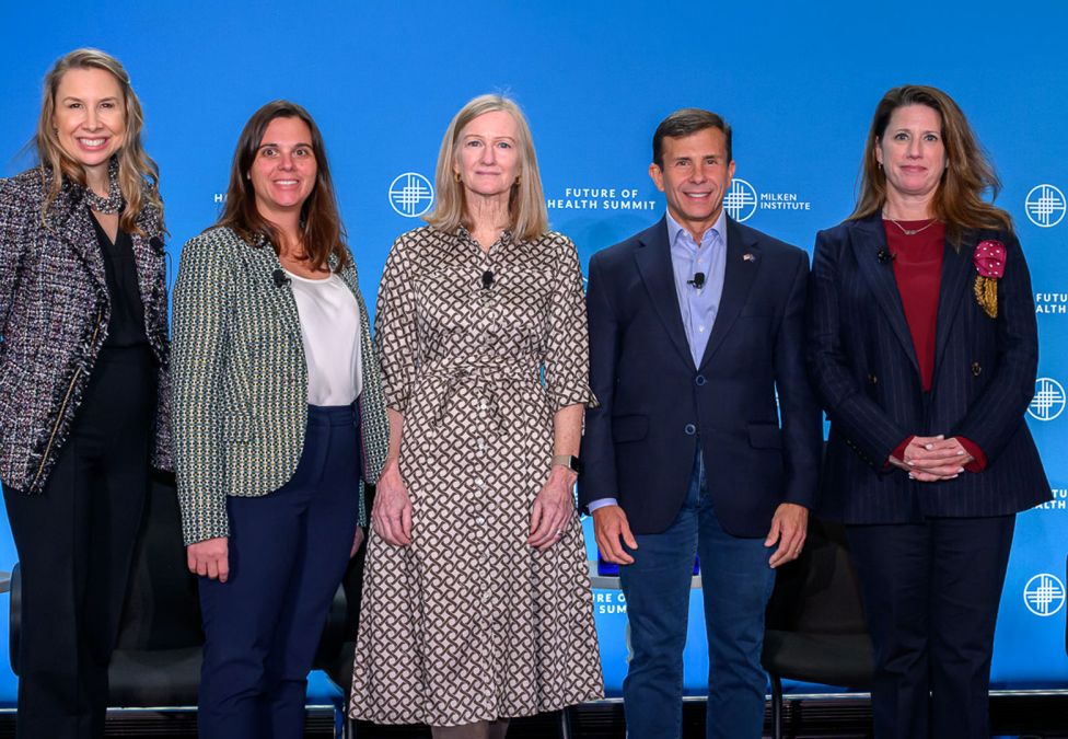 Five panelists stand on stage in front of a blue Milken Institute Future of Health Summit backdrop, smiling at the camera.