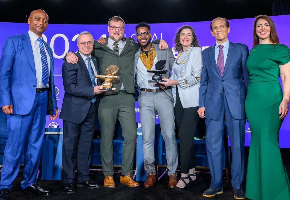 A group of seven people stand together on stage at a Milken Institute event, smiling and holding awards. The backdrop features the Milken Institute logo and event branding in blue and purple tones.
