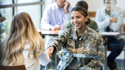An image of a woman with white skin and brown hair in a bun, wearing a military uniform and shaking hands with a blonde woman with white skin. The image leads into an article about reinvigorating the career pathways for veterans.