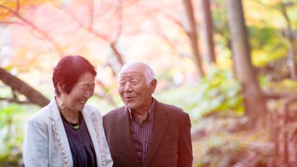 An image of an elderly man and woman who are Asian in appearance with white skin, the woman has short black hair and the man has short white hair. They are walking on a trail underneath trees.