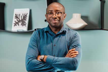 An image of a man named Ola Oyetayo who is the founder of a company called Verto, a winner of the Milken-Motsepe Prize. He has a bald head, brown skin, and a slight beard and is wearing round glasses, a blue collared shirt, and standing in front of a blue decorative background with crossed arms and slight smile. 