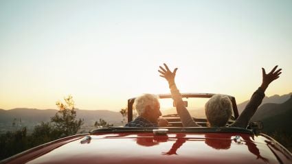 An image of two people's silhouettes in a red convertible, driving into the sunset with one of them holding his or her hands in the air.