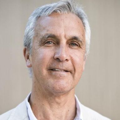 An image of a man named Michael Hodin. He is older in appearance and has short white hair and white skin. He is wearing a white shirt and standing against a beige background.