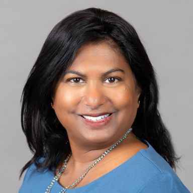 An image of a female-presenting person who is Indian in appearance. She is smiling at the camera and standing against a gray background. She has long black hair, tan skin, brown eyes, and is wearing a light blue shirt with a long necklace.