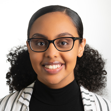 An image of a female-presenting person named Hermela Assefa. This person has light brown skin and dark brown curly hair that is smooth on the top and curly on the bottom. This person is wearing black framed glasses and a striped black and white blazer over a black shirt while smiling at the camera against a white background.