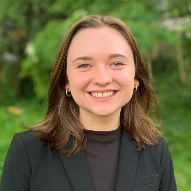 An image of a woman named Lillian Parr. She is standing against a green outdoor background and smiling at the camera. She has light white skin, shoulder-length brunette hair, and is wearing a black blazer over a black shirt.