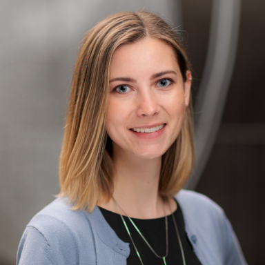 An image of a woman named Samantha Shumm. She is standing against a blurred gray background and smiling at the camera. She has light white skin, shoulder-length straight brownish blonde hair, and blue eyes. She is wearing a light blue cardigan over a black shirt.