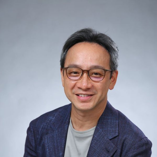 A headshot of a man who is Asian in appearance with short gray and black hair, and round reddish glasses. He is wearing a dark blue blazer and a gray shirt. 