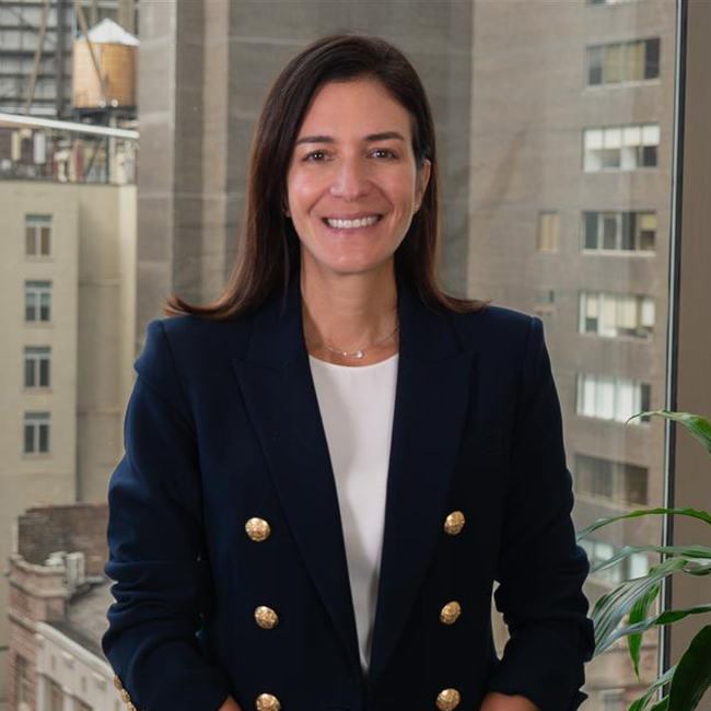 An image of a woman named Vanessa Dager. She is standing against a city building background and smiling at the camera. She has white skin and shoulder-length brown hair, and she is wearing a blue blazer with gold buttons over a white shirt.