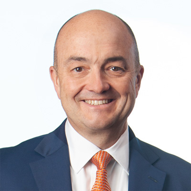 A headshot of a man named Andrew Sullivan. He has white skin, a bald head, and is wearing a blue suit with an orange tie while smiling at the camera against a white background.