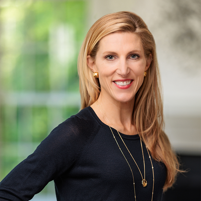 An image of a woman named Vanessa Kerry. She is standing against an ambiguous background with her hands on her hips and smiling at the camera. She has long blonde hair, white skin, and is wearing gold stud earrings and a dark blue long-sleeved shirt with two gold long-length necklaces.
