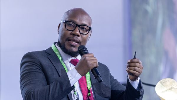 A speaker at the Milken-Motsepe Prize in FinTech event holds a microphone and clicker while presenting on stage. He wears glasses, a dark suit, a white shirt, and a red tie, with a green "SPEAKER" lanyard around his neck.