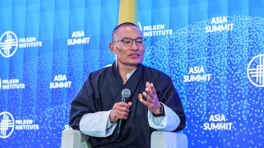 An image of a man mid-speech sitting on stage and speaking at the Milken Institute's Asia Summit 2024 against a blue Milken Institute backdrop. He is the Prime Minister of Bhutan, and he is Asian in appearance with tan white skin, buzzed black hair, and is wearing round glasses and dark blue formal robe-like attire.