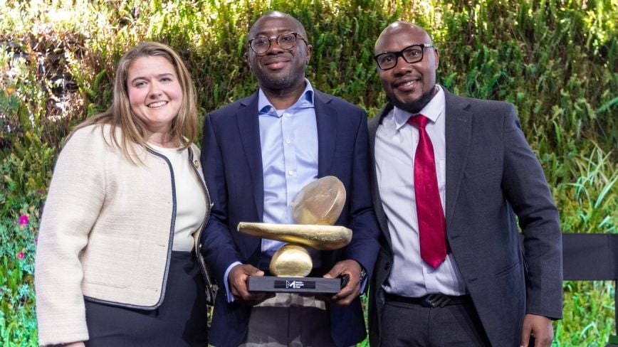 Three individuals stand together smiling in front of a lush green backdrop. The person in the center holds a golden sculptural trophy with a base labeled “Milken-Motsepe Prize.” This image depicts the award presentation for the Milken-Motsepe Prize in FinTech, celebrating innovation in financial technology.