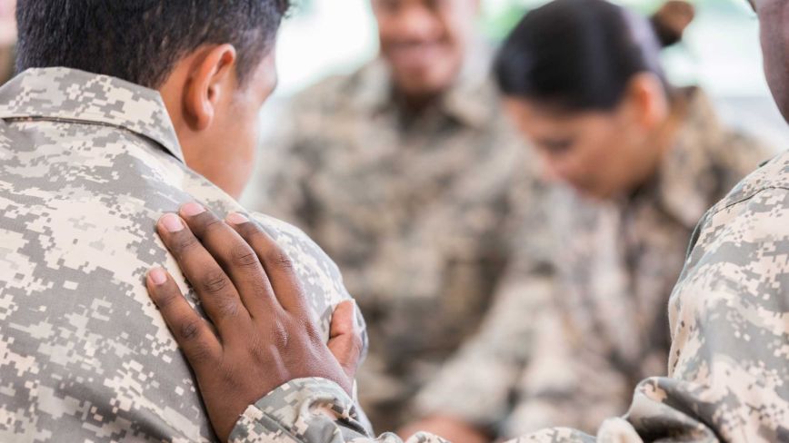An up close image of a person putting a hand on another person's back. They are wearing camouflage fatigues and there are other people wearing matching fatigues blurred in the background.