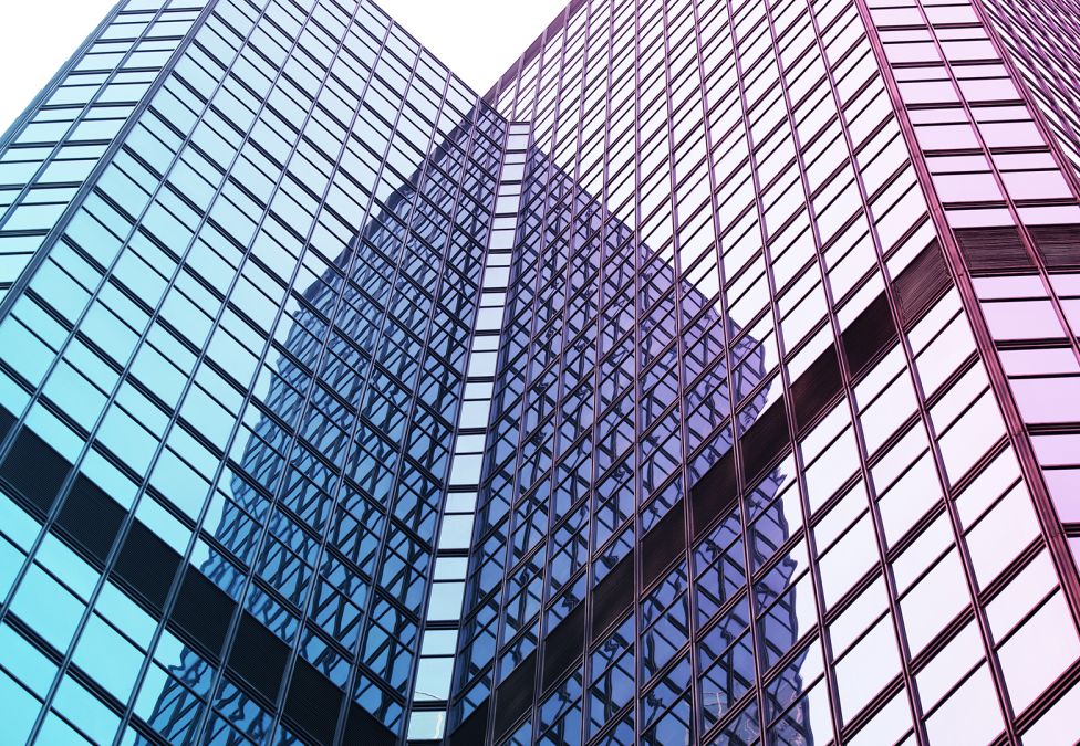 An image of a skyscraper building, upclose, pointing upwards and showing its glass windows with blue-ish and purple-ish hues reflected off of the windows.