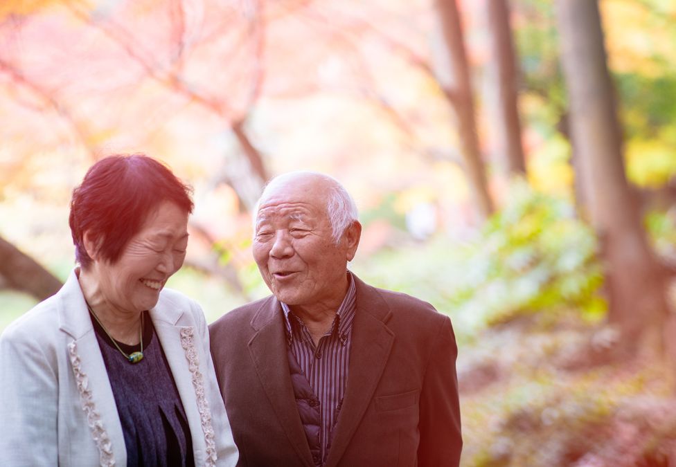 An image of an elderly man and woman who are Asian in appearance with white skin, the woman has short black hair and the man has short white hair. They are walking on a trail underneath trees.