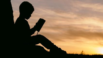 A child's silhouette sitting down reading a book against a sunset
