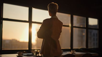 A person, photographed from behind, stands in an office and looks out the window as the sun sets.