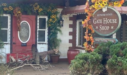 An image of the outside of the Biltmore Estate's Gate House Shop. The building features a red door, surrounded by flowers, and shows a knocked over table and chairs due to the aftermath of hurricane activity. 