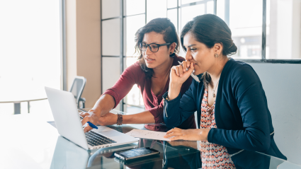 A photo of two Latin American women with dark hair and professional attire sitting in an office and looking at a shared laptop.
