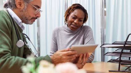 An image of a woman with brown skin and short brown braids, wearing a grey sweater, talking to a male doctor with grey hair, a gray beard, white skin, glasses, a green sweater, and wearing a stethoscope. They are reviewing a tablet together and the image leads to a Milken Institute article around a preventative health roundtable. 